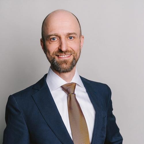  A smiling bald man with a beard and blue eyes wears a dark blue suit, a white shirt, and a gold and brown patterned tie, posing for a headshot against a plain light gray background.